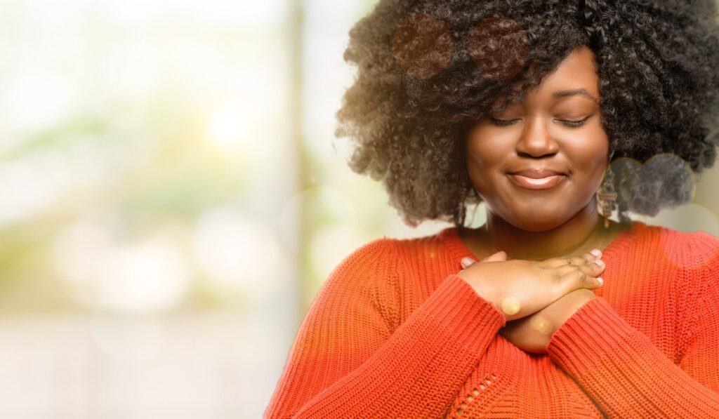 An African-American woman clasps her hands to her chest looking content with herself.